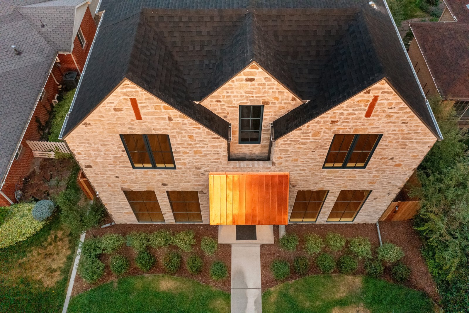 Aerial view of a modern two-story stone house with a dark roof, large symmetrical windows, and a striking copper awning over the front entrance. The house is surrounded by a neatly landscaped yard with shrubs and a walkway leading to the front door.